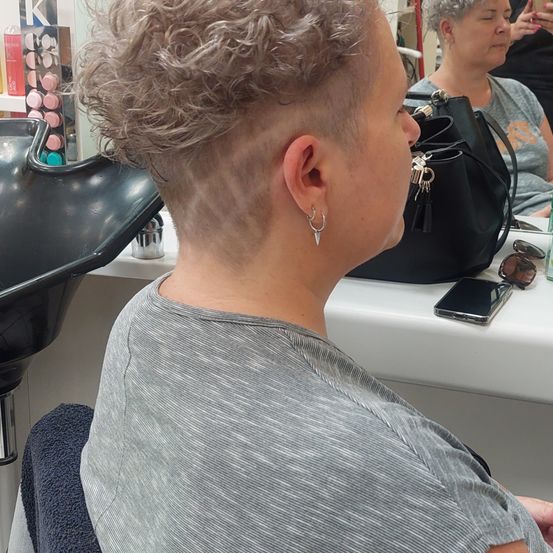 Eine Frau mit kurzen, lockigen Haaren sitzt in einem Salon und schaut in einen Spiegel. Hinter ihr eine Frau mit Handtasche und Handy. Der Salon hat eine weiße Theke mit verschiedenen Gegenständen.