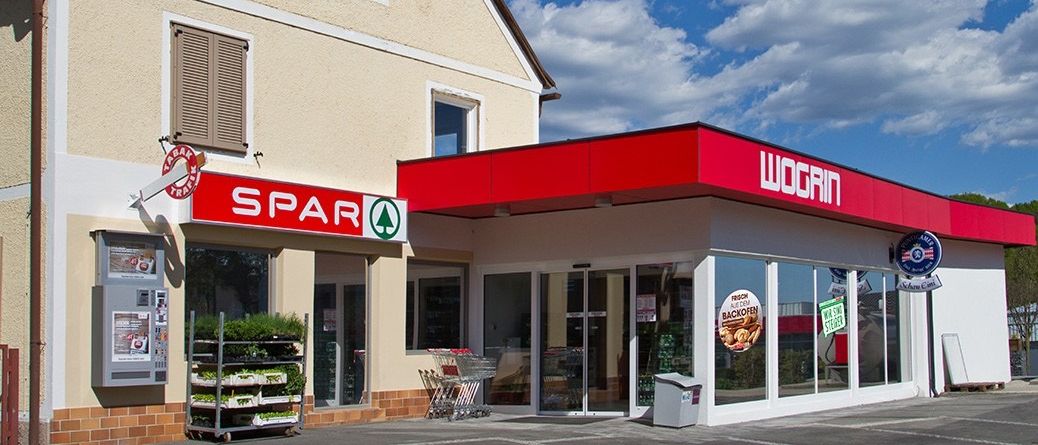 Ein Spar-Supermarkt steht neben einer Wogrin-Tankstelle. Beide haben Glasfenster und rote Dächer. Ein Pflanzenständer steht vor dem Supermarkt. Der Himmel ist blau mit einigen Wolken.