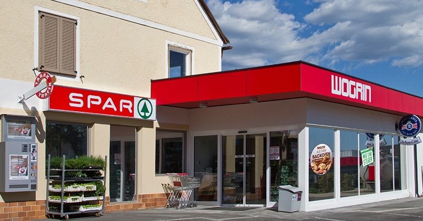 Ein Spar-Supermarkt steht neben einer Wogrin-Tankstelle. Beide haben Glasfenster und rote Dächer. Ein Pflanzenständer steht vor dem Supermarkt. Der Himmel ist blau mit einigen Wolken.