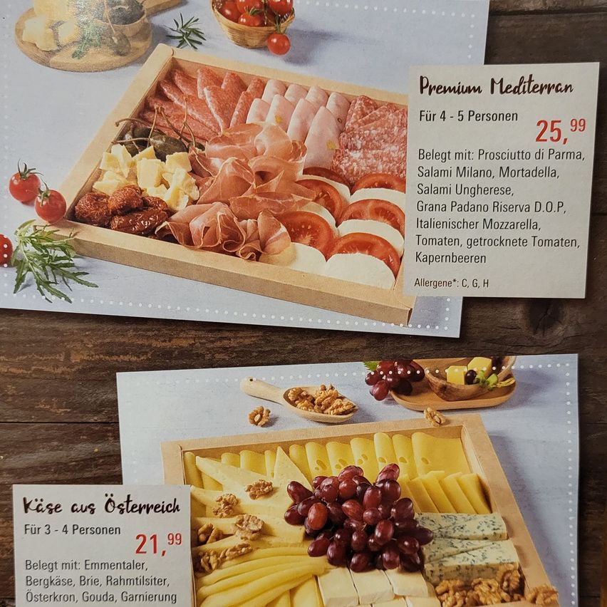 Das Bild zeigt zwei Holzschalen mit verschiedenen Käse- und Fleischsorten. Die obere Schale enthält eine Auswahl an Fleisch und Käse, darunter Prosciutto, Salami und Mozzarella, zum Preis von 25,99 Euro für 4-5 Personen. Die untere Schale ist eine Käseplatte mit Trauben, Walnüssen und verschiedenen Käsesorten, zum Preis von 21,99 Euro für 3-4 Personen.