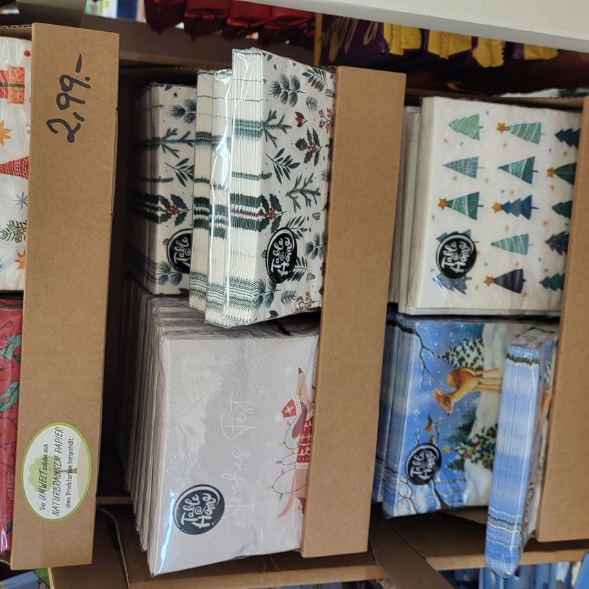 Ein Regal in einem Geschäft zeigt mehrere Kartons mit weihnachtlich bedruckten Servietten. Die Kartons sind aus Pappe und enthalten jeweils mehrere Servietten. Die Servietten haben verschiedene weihnachtliche Motive, darunter Bäume, Sterne und ein Reh. Der Preis der Kartons ist auf der linken Seite angegeben.