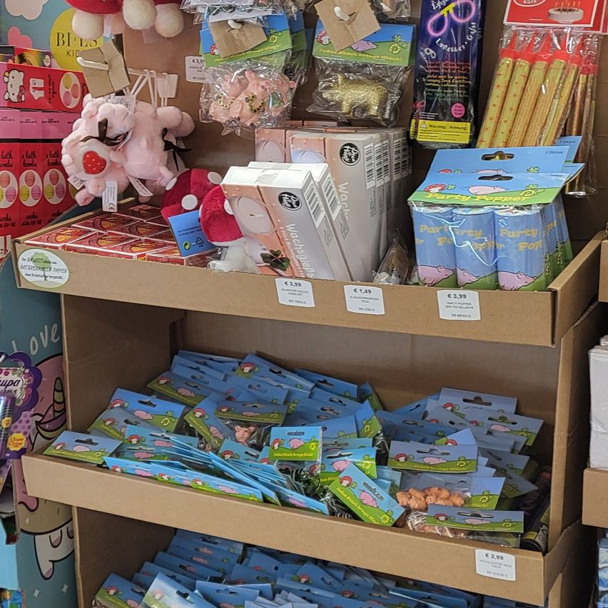 Ein Ladenregal zeigt eine Vielzahl von Artikeln, darunter Plüschtiere, Partyzubehör und verschiedene farbenfrohe Packungen mit Süßigkeiten und Snacks, mit sichtbaren Preisschildern.