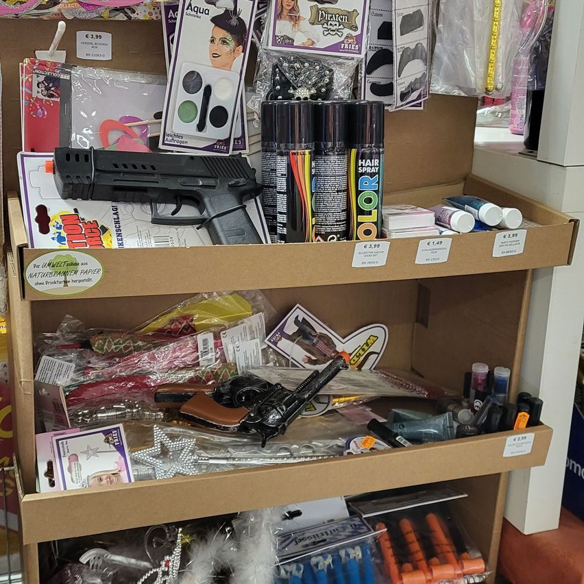 Ein braunes Kartonregal zeigt verschiedene Gegenstände, darunter eine schwarze Spielzeugpistole, Farbsprühdosen und diverse Zubehörteile. Das Regal enthält bunte Objekte und einige sind mit Preisen versehen.