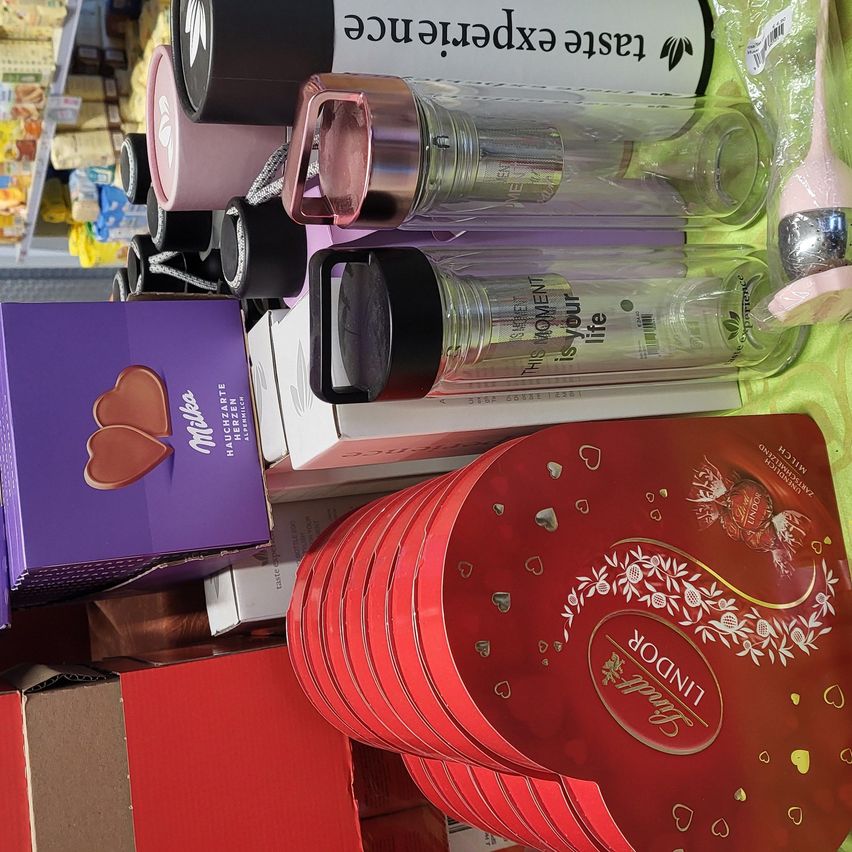 Verschiedene Produkte sind ausgestellt, darunter Thermosflaschen, Schokoladen und Kartons. Die Thermosflaschen haben Texte wie 'Dieser Moment ist dein Leben.' Die Schokoladen sind in lila und roten Schachteln. Einige Schachteln haben das Wort 'Lindt' darauf.