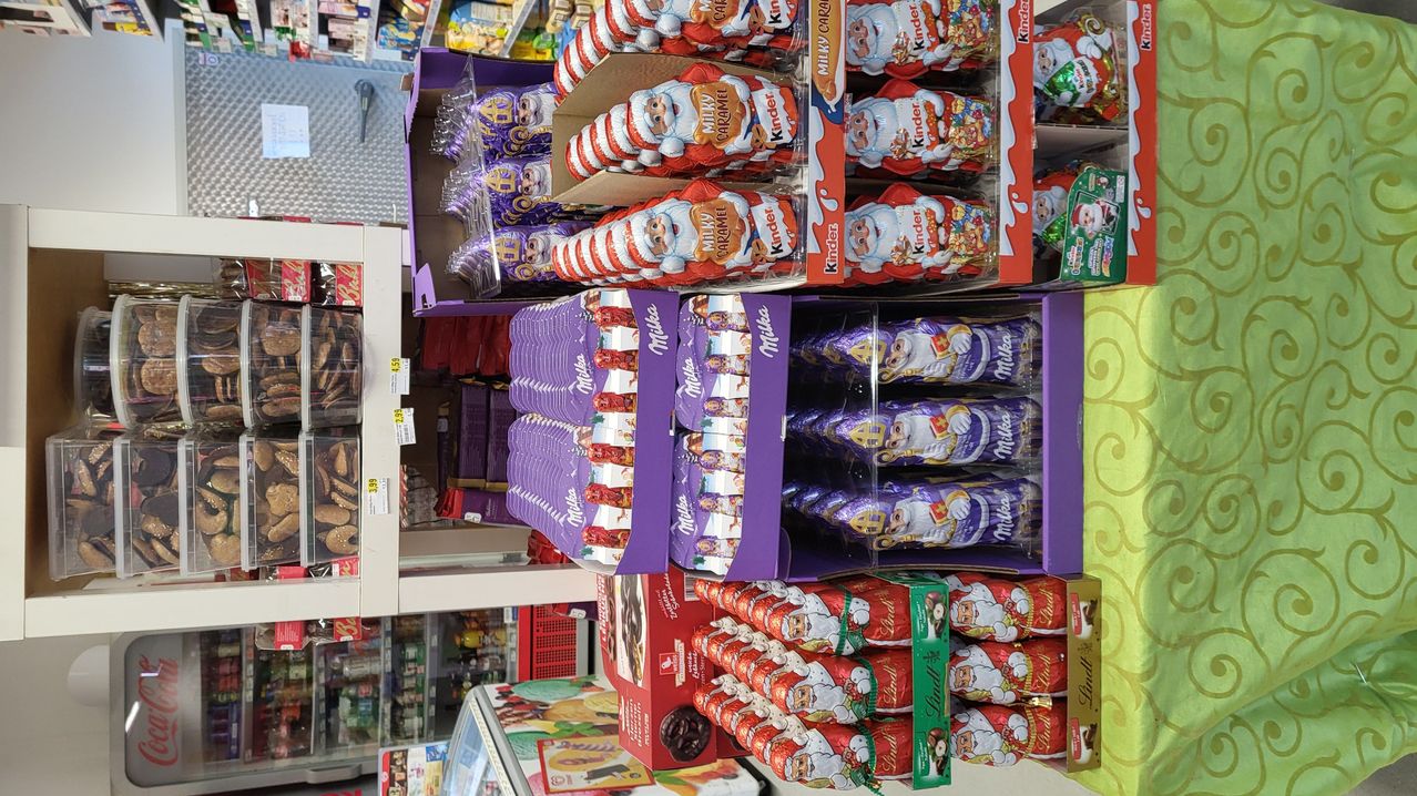 A store displays various gift boxes featuring Santa Claus, including Kinder Surprise, Milka, and Lindt chocolates. The boxes are in red, purple, and green colors, arranged in neat rows on shelves.