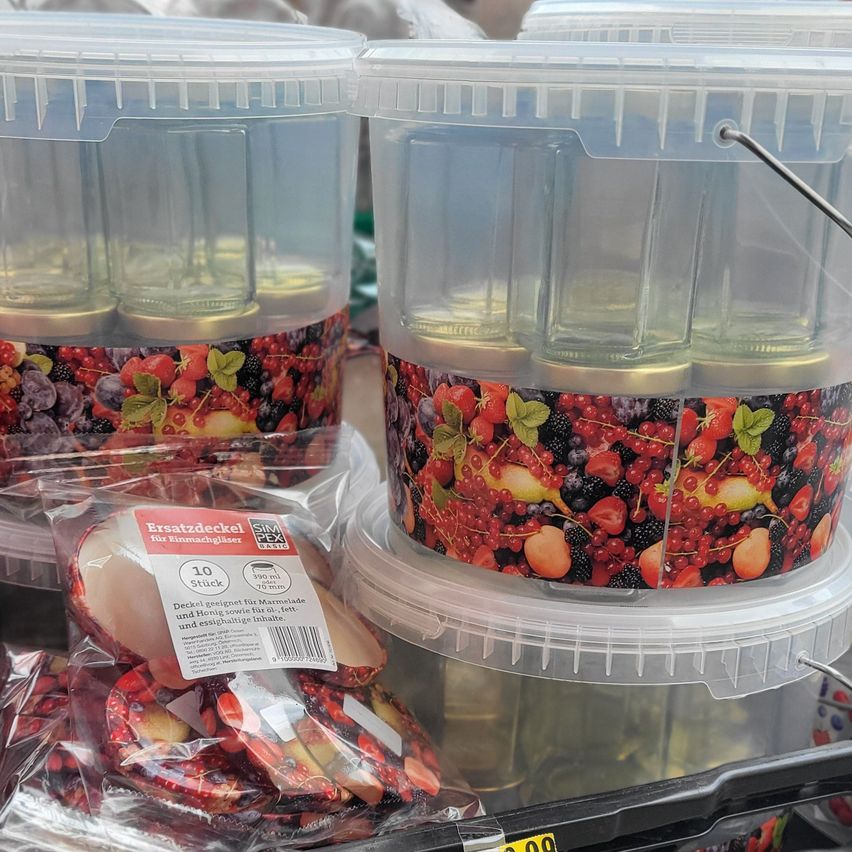 Several clear plastic containers with lids, each containing glass jars with lids and fruit-themed designs. Some containers have labels with text and numbers. One container has a handle and a barcode.