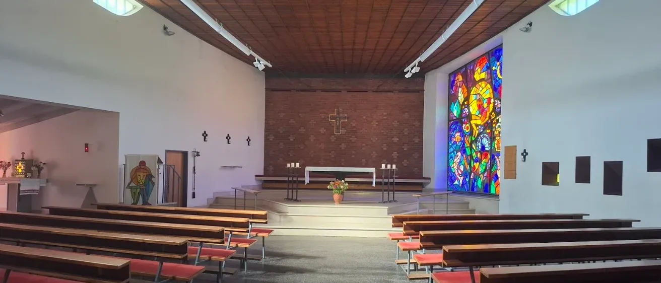 Ein leerer Kircheninnenraum mit hölzernen Kirchenbänken, einem farbenfrohen Buntglasfenster und einem schlichten Altar.