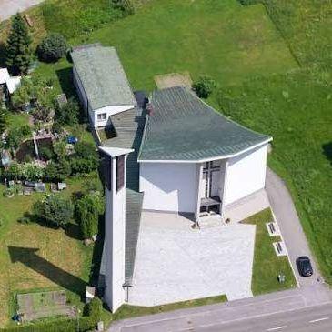 Luftaufnahme eines modernen weißen Gebäudes mit grauem Dach, umgeben von grünem Gras und Bäumen, mit einer Einfahrt, die zu einer Straße führt.