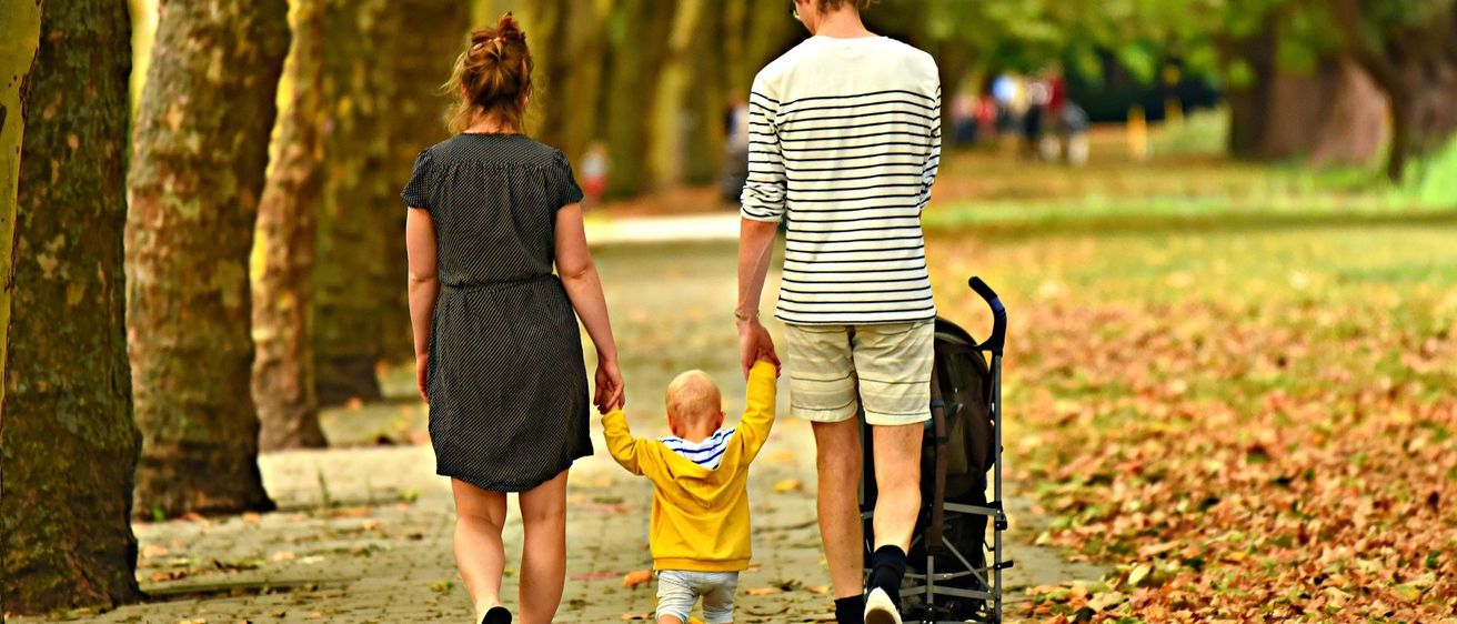 Eine vierköpfige Familie geht auf einem gepflasterten Weg in einem Park. Die Mutter und der Vater halten die Hände des Kindes, während der Vater einen Kinderwagen schiebt.