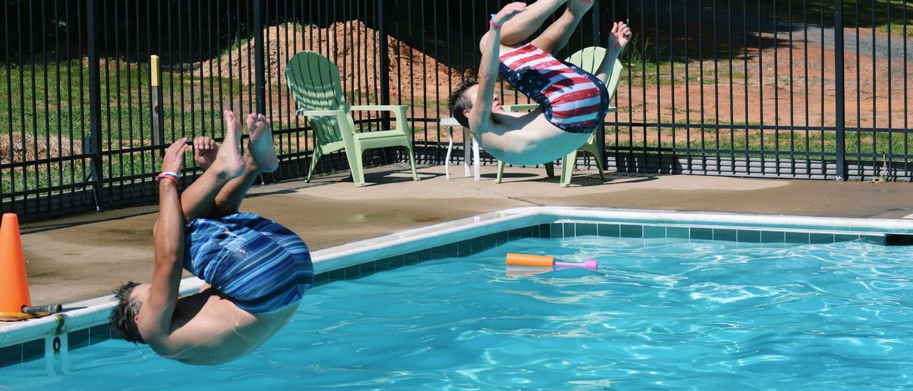 Zwei Männer springen in einen Pool. Ein Mann trägt einen blau-weiß gestreiften Badeanzug. Im Hintergrund befinden sich grüne Stühle und ein schwarzer Zaun.