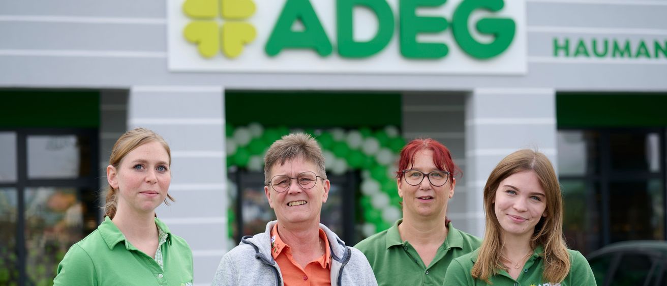 Vier Frauen stehen vor einem Laden mit dem Schild 'ADEG' über ihnen. Sie tragen grüne Poloshirts, wobei die mittlere Frau ein orangefarbenes Shirt und eine Brille trägt.