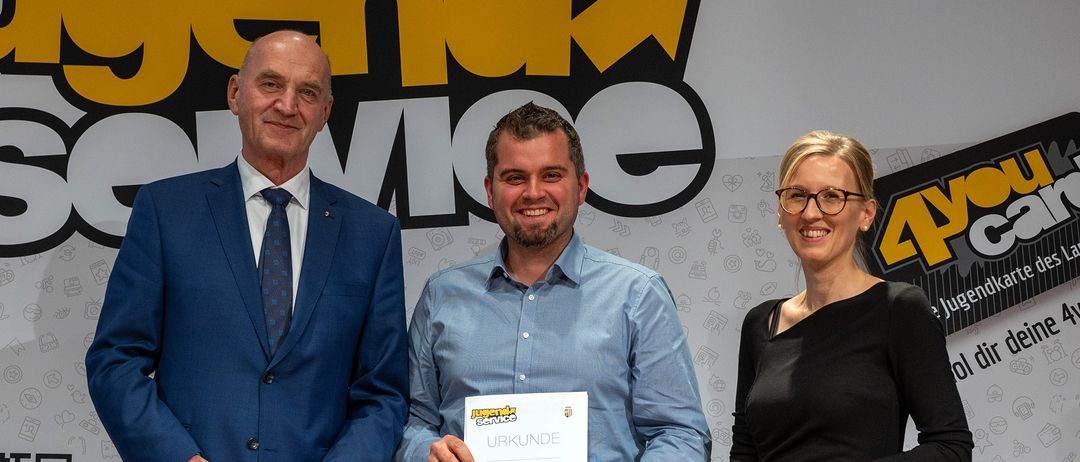 Drei Personen stehen vor einem Hintergrund. Der Mann in der Mitte hält ein Zertifikat. Der Mann auf der linken Seite trägt einen blauen Anzug. Die Frau auf der rechten Seite trägt eine Brille.