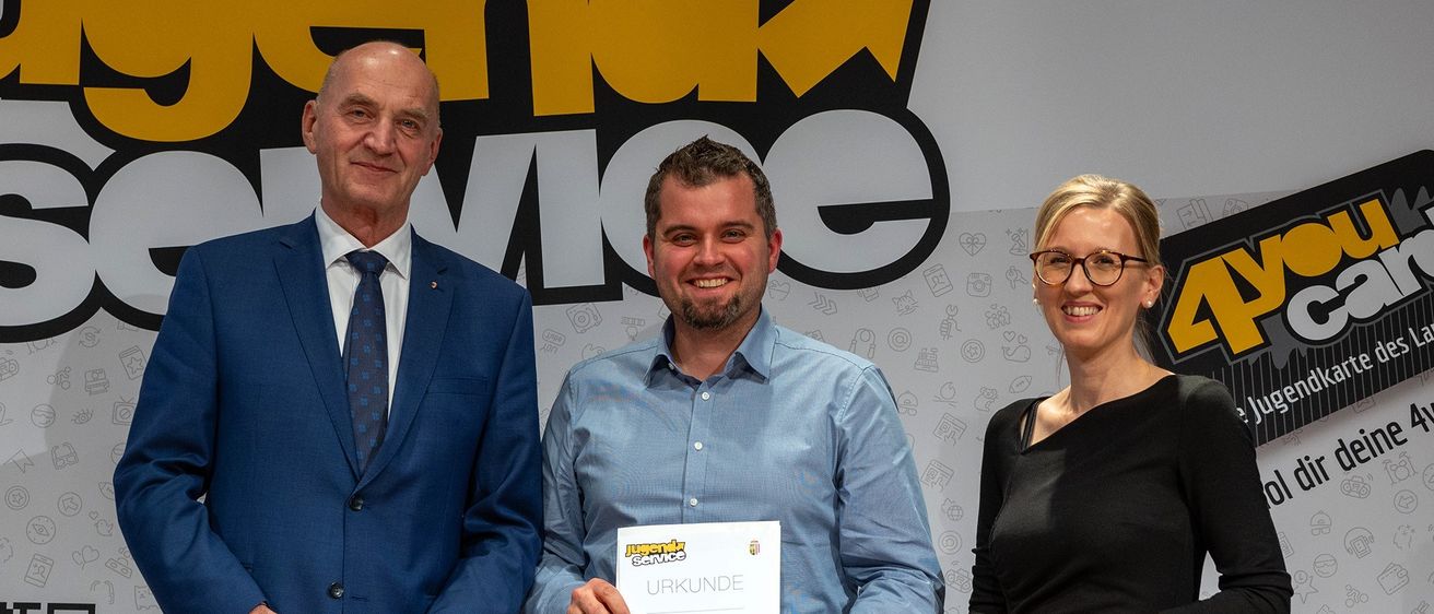 Drei Personen stehen vor einem Hintergrund. Der Mann in der Mitte hält ein Zertifikat. Der Mann auf der linken Seite trägt einen blauen Anzug. Die Frau auf der rechten Seite trägt eine Brille.