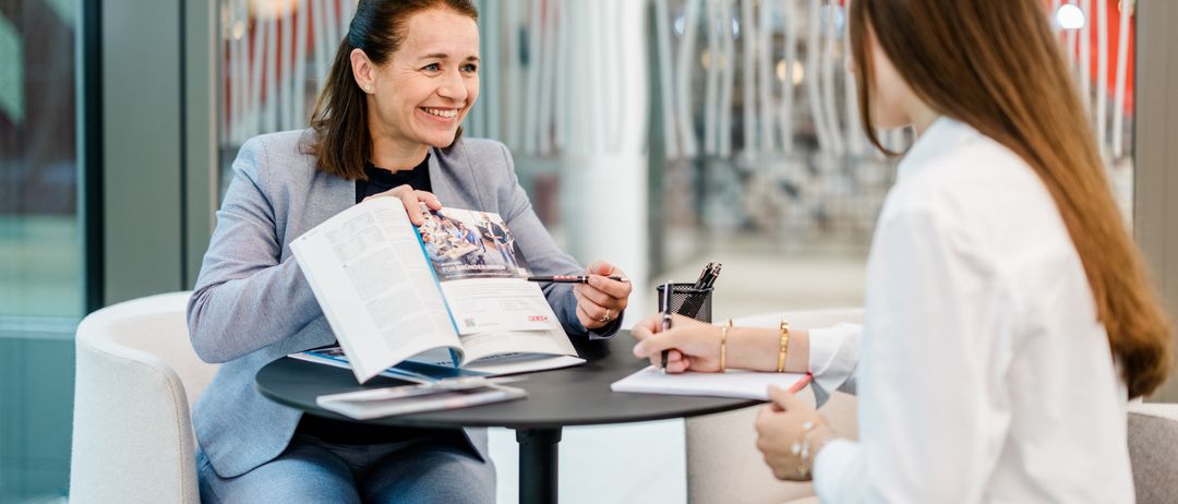 Zwei Frauen sitzen sich gegenüber an einem Tisch. Die Frau in einem Blazer hält eine Broschüre und einen Stift und lächelt. Die andere hält einen Stift und Papier und führt ein Gespräch.