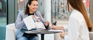 Zwei Frauen sitzen sich gegenüber an einem Tisch. Die Frau in einem Blazer hält eine Broschüre und einen Stift und lächelt. Die andere hält einen Stift und Papier und führt ein Gespräch.