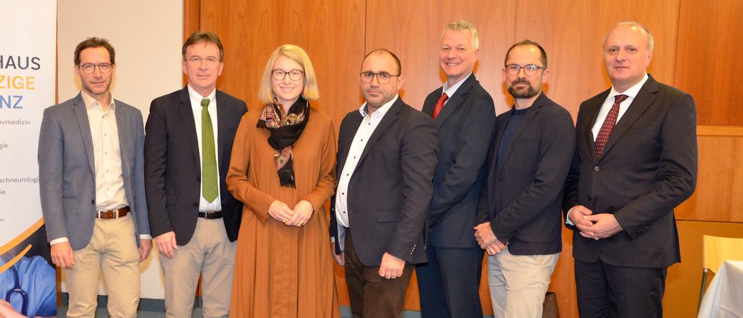 Eine Gruppe formell gekleideter Personen steht gemeinsam in einem Raum, lächelt und posiert für ein Foto vor einer Holzwand.