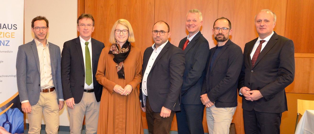 Eine Gruppe formell gekleideter Personen steht gemeinsam in einem Raum, lächelt und posiert für ein Foto vor einer Holzwand.