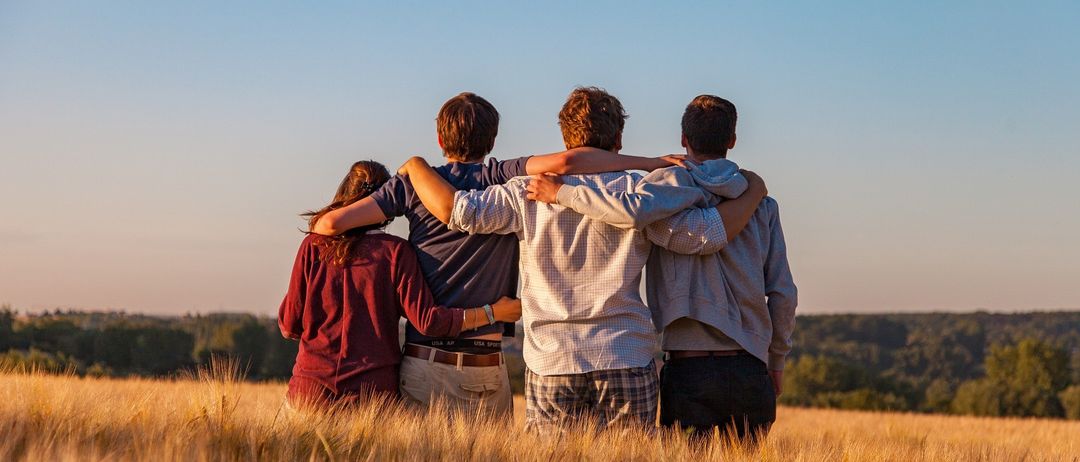 Vier Personen stehen in einem Weizenfeld mit den Armen umeinander geschlungen und wenden sich von der Kamera ab. Sie stehen an einem klaren Tag mit blauem Himmel.