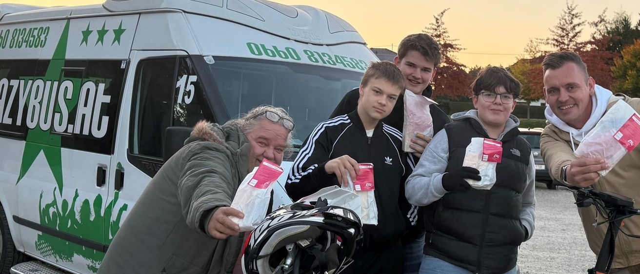 Fünf Personen stehen neben einem Van und einem Motorrad, lächeln und halten rote Pakete. Auf dem Van steht 'y6us.at'. Das Motorrad ist schwarz und hat einen Helm auf dem Sitz. Der Boden ist mit Kies bedeckt.