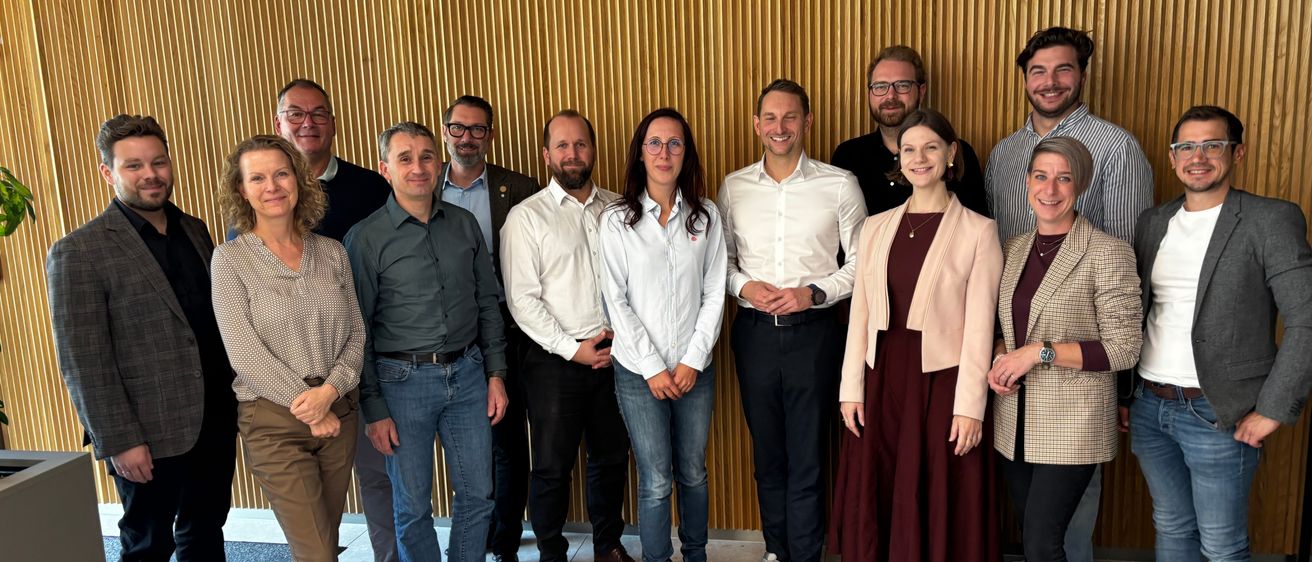 Eine Gruppe von Menschen in Geschäftskleidung steht vor einer goldenen Wand. Sie scheinen für ein Foto zu posieren. Sie alle sehen glücklich aus und lächeln.