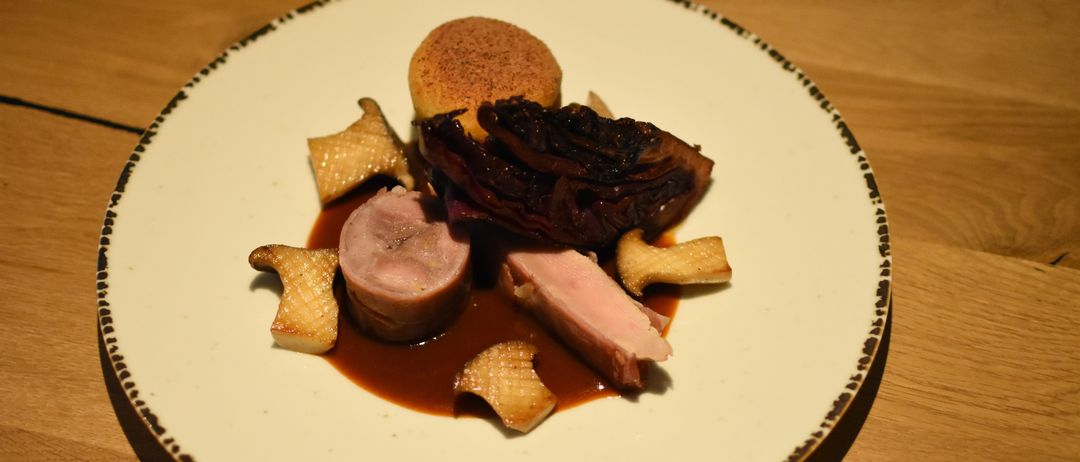 Ein weißer Teller mit schwarzem Rand enthält ein anspruchsvolles Gericht. Es besteht aus zwei Schweinefleischscheiben, einem kleinen Stück Brot und einer Portion dunkelroten Krauts, alles bedeckt mit brauner Soße.