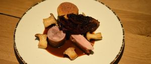 Ein weißer Teller mit schwarzem Rand enthält ein anspruchsvolles Gericht. Es besteht aus zwei Schweinefleischscheiben, einem kleinen Stück Brot und einer Portion dunkelroten Krauts, alles bedeckt mit brauner Soße.