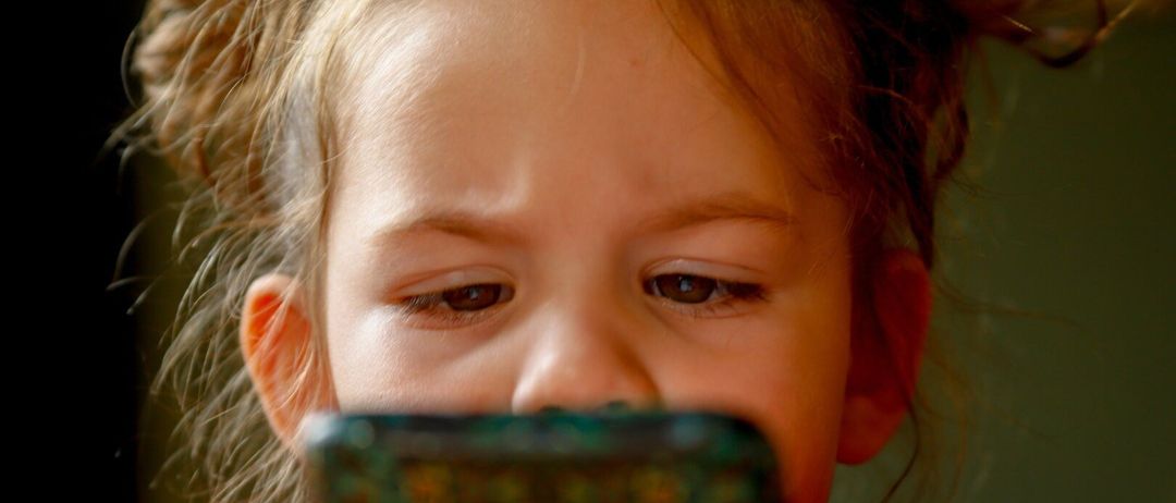 Ein kleines Mädchen mit blonden Haaren hält ein grünes Smartphone und schaut auf den Bildschirm, möglicherweise konzentriert auf eine Nachricht.