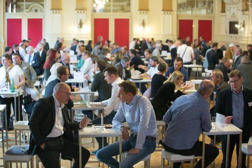 Eine große Gruppe von Menschen sitzt in einem Konferenzsaal an Tischen und führt Gespräche. Der Raum ist gut beleuchtet, mit roten Vorhängen und Kronleuchtern.
