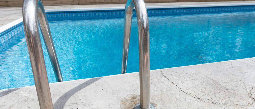 An outdoor swimming pool with clear blue water and stainless steel handrails on the pool deck. The pool has a wooden fence surrounding it.