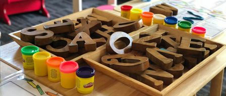 Ein Holztablett mit Buchstaben des Alphabets und Play-Doh-Behältern auf einem Tisch. Ein roter Stuhl und Klavier im Hintergrund.