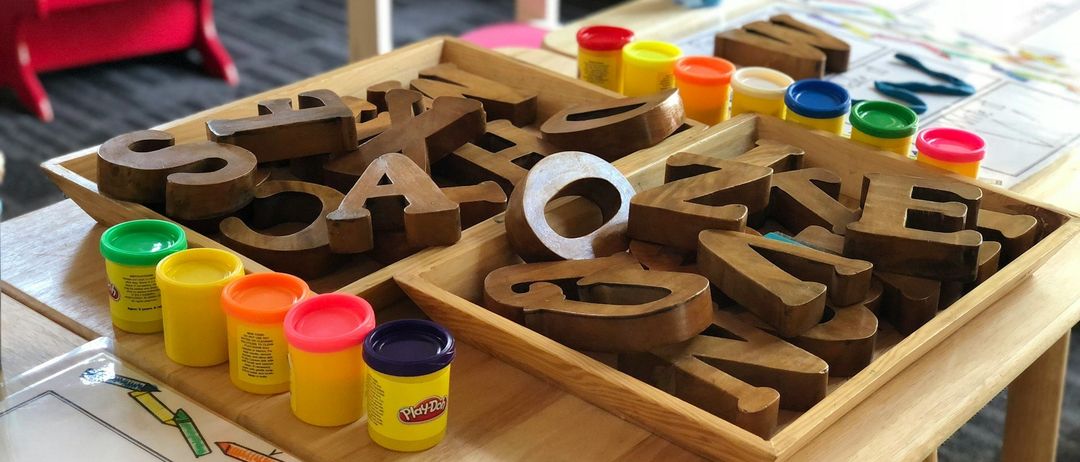Ein Holztablett mit Buchstaben des Alphabets und Play-Doh-Behältern auf einem Tisch. Ein roter Stuhl und Klavier im Hintergrund.