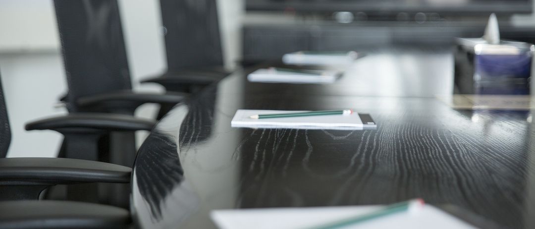 Ein leerer Konferenztisch mit Stiften und Notizblöcken darauf, mit schwarzen Lederstühlen im Hintergrund.