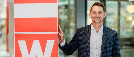 Ein Mann steht neben einem großen orangefarbenen Schild mit den Buchstaben WKO in weiß. Er trägt einen dunkelblauen karierten Anzug und ein weißes Hemd. Er lächelt und scheint für ein Foto zu posieren.