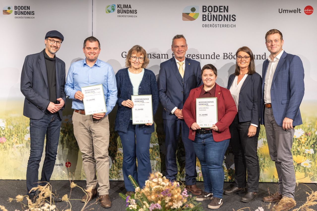Eine Gruppe von Personen mit Urkunden steht vor einem Hintergrund. Eine Frau hält eine Urkunde für 'Bodenbundnis Gemeinde Kirchberg-Theining'. Ein Schild hinter ihnen lautet 'Klima Bundnis Oberosterreich Boden Bundnis Oberosterreich'.