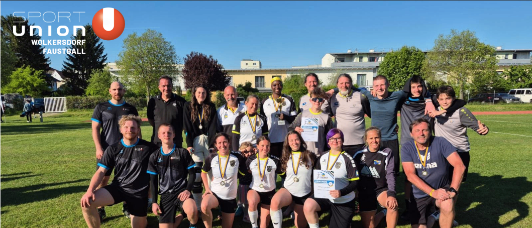 Eine Gruppe von Menschen, möglicherweise ein Sportteam, posiert für ein Foto auf einem Rasenfeld. Sie tragen Medaillen und halten Urkunden. Hinter ihnen stehen Bäume und Gebäude.