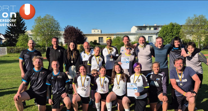 Eine Gruppe von Menschen, möglicherweise ein Sportteam, posiert für ein Foto auf einem Rasenfeld. Sie tragen Medaillen und halten Urkunden. Hinter ihnen stehen Bäume und Gebäude.