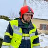 Zwei Feuerwehrleute in gelben und schwarzen Uniformen stehen im Schnee neben einem Feuerwehrauto. Einer hat einen roten Helm, der andere einen gelben Helm mit der Aufschrift 'BRUHLIGEN'. Ein Haus mit Schnee auf dem Dach steht hinter ihnen.