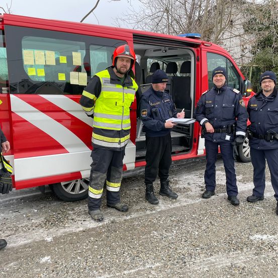 Vier Männer stehen vor einem roten Van. Drei tragen Polizeiuniformen, einer eine reflektierende gelbe und schwarze Weste. Ein Polizist schreibt auf ein Papier.