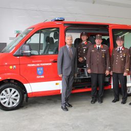 Vier Personen, darunter ein Mann in Anzug und drei in Militäruniformen, stehen neben einem roten Feuerwehrwagen mit Freiwillige Feuerwehr Feiting auf der Seite.