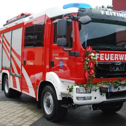 Ein roter Feuerwehrwagen mit weißen Akzenten ist draußen geparkt. Die Vorderseite hat Blumen und das Wort 'Feuerwehr'. Das Kennzeichen lautet 'LBV51FF'.