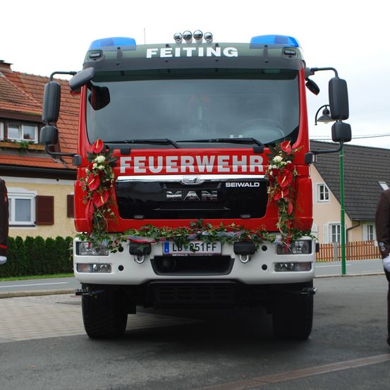 Ein roter Feuerwehrwagen mit der Aufschrift 'Feuerwehr' steht vor einem Gebäude. Er hat Blumen an der Front und ein Nummernschild.