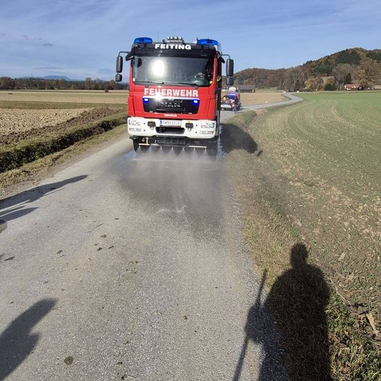 Ein roter Feuerwehrwagen fährt auf einer Straße mit einem vorbeigehenden Menschen und hinterlässt eine Wasserspritze. Ein Auto folgt dahinter.