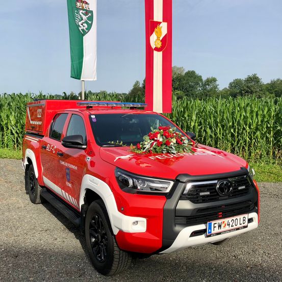 Ein roter Toyota-Pick-up, geschmückt mit Blumen und einem Kranz, steht in einem Maisfeld mit Flaggen im Hintergrund.