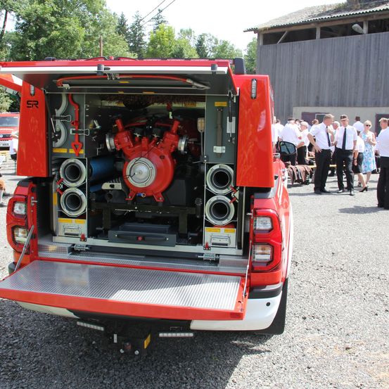 Ein rotes Fahrzeug mit offenem Heck, das eine große rote Feuerpumpe, Schläuche und andere Feuerwehrausrüstung zeigt. Hinter ihm steht eine Gruppe von Menschen in formeller Kleidung.