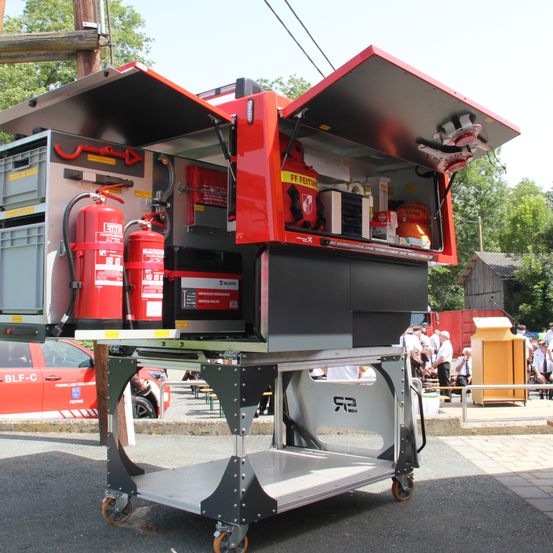 Ein geöffneter Ausrüstungseinheit einer Feuerwache mit Feuerlöschern und Aufbewahrungsboxen. Ein rotes Auto und mehrere Personen im Hintergrund.