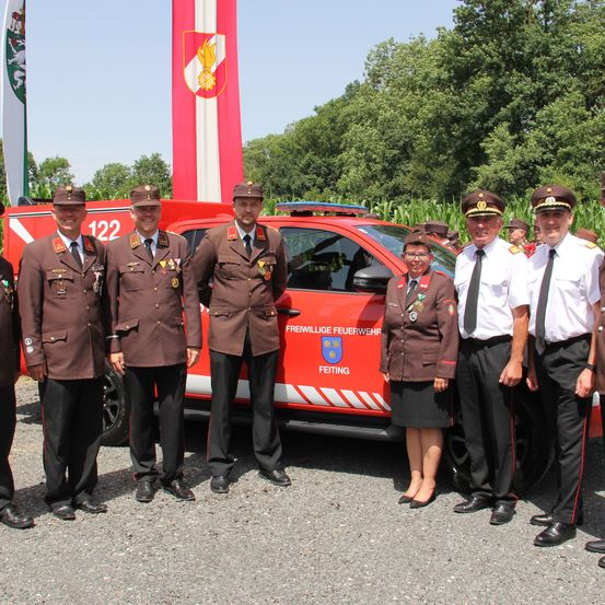 Eine Gruppe von Feuerwehrleuten steht vor einem roten Feuerwehrauto. Sie tragen Uniformen und einige von ihnen tragen Medaillen. Hinter ihnen sind Bäume und ein Pfahl.