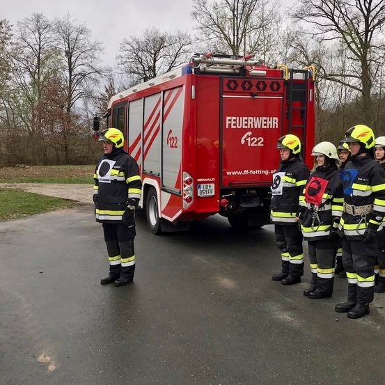Eine Gruppe von Feuerwehrleuten steht in Formation vor einem Feuerwehrauto auf einer asphaltierten Straße, mit Bäumen im Hintergrund.