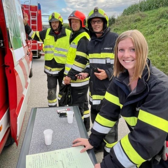 Drei Feuerwehrleute in gelben und schwarzen Uniformen posieren für ein Foto mit einer lächelnden Frau vor einem Tisch mit Bechern und einem Papier. Ein roter Krankenwagen ist hinter ihnen geparkt.