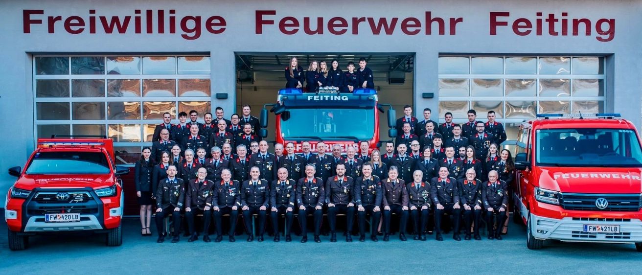 Ein Gruppenfoto einer Feuerwehrmannschaft, bei der mehrere Feuerwehrleute vor einem Feuerwehrwagen stehen, während andere in Reihen darunter sitzen.