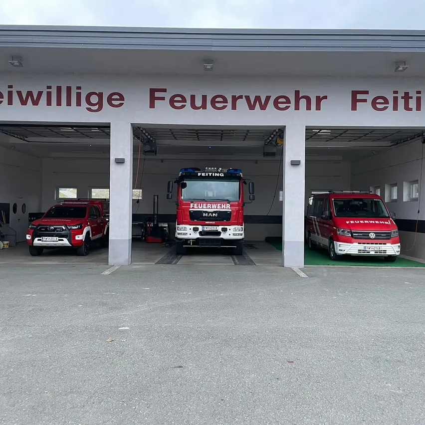 Eine Feuerwehrstation mit dem Namen Freiwillige Feuerwehr Feiti ist zu sehen. Drei rote Fahrzeuge sind drinnen geparkt, darunter ein LKW und zwei Autos.